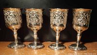 Front view of four ornate vintage footed goblets with detailed embossed designs, showing the Hollywood Regency style and scuffed plating on a wooden surface.