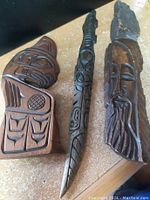 Three indigenous wood carvings displayed on a surface: a mask, a slender staff, and a bust figure.