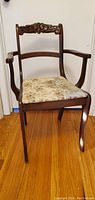 Front view of one antique wooden armchair showing carved backrest and floral upholstered padded seat on hardwood floor.