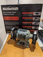 Delta 6-inch bench grinder with a Mastercraft bench grinder stand box in background placed on a wooden surface.