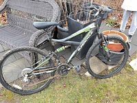 Photo of a black Supercycle mountain bike leaning against a black wicker chair outdoors, showing the 18-inch frame, 26-inch wheels, padded seat, gear system, and front suspension fork.