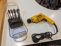 Yellow DeWalt corded drill with black grip and power cord coiled beside a clear plastic case containing multiple spade drill bits