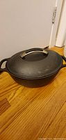 Photo showing KitchenAid black cast iron Dutch oven with lid on wooden floor, highlighting the oval shape, side handles, and lid with metal handle.