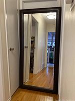 Front view of the full-length mirror standing against a wall showing the brown faux leather frame and clear mirror surface reflecting a hallway.
