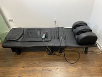 Side view of black collapsible massage bed showing full length, faux leather surface, headrest, remote control resting on surface, and power cable hanging below.