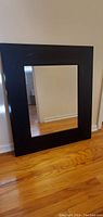 Medium mirror with brown wooden frame leaning against white wall on wooden floor