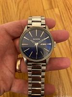 Watch held in hand, showing blue dial and stainless steel bracelet.