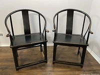 Pair of black wooden chairs styled after 17th century Ming Dynasty design with rounded backs and armrests, placed side by side on a wooden floor.