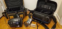 Wide shot showing both video cameras, their carrying bags, and accessories on a flat surface.