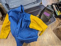 Blue and yellow protective welding coat laid open next to auto-darkening welding helmet in plastic bin