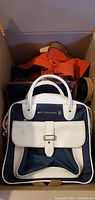 Photo of a box containing various purses and bags including a white and navy blue Air Canada handbag with buckle front pocket, partially visible orange bags behind.
