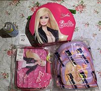 Photo showing a pink Barbie cosmetic case, a pink Barbie lunch box, and a small colorful backpack on floral background alongside a soda can for scale.