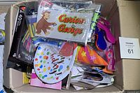 Box filled with assorted birthday party supplies including a Curious George birthday banner, colorful party blowers, candles, napkins, and plates.