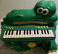 Front view of the green crocodile-shaped children's piano keyboard with all keys and buttons visible, showing overall appearance and condition.