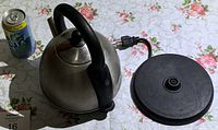 Stainless steel electric kettle and black electric base with cord and plug beside a can for scale