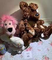 Photo showing four different plush toys on a floral fabric background, including a large brown bear holding smaller bears, a pink plush with fuzzy hair, a white plush, and a smaller brown plush toy.