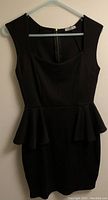 Full view of black sleeveless peplum dress on hanger against wall.