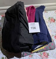 Stack of various women's pants and clothing, mostly in dark colors with some bright pink and blue items.