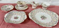 Photo showing the full selection of bone china pieces arranged on a cloth surface: teacup, saucers, creamer, and plates with floral and ribbon detail in Bridal Gown pattern.