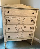 Front view showing the four drawers with black painted decorative designs on middle three drawers and metal ring handles, along with turned legs and distressed white finish.