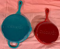 Set of two Crock-Pot Artisan cast iron skillets, one teal and one red enamel, laying on a pink fabric surface showing bottom views with embossed brand name.