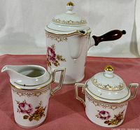 Full coffee set showing coffee pot with wooden handle, creamer, and lidded sugar bowl with floral and gold decorative pattern on white porcelain