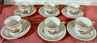 Wide photo showing six demitasse cups and six matching saucers set on a pink fabric background.