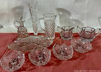 Photo showing nine vintage clear glass pressed and cut pieces including decanter, vase, pitcher, serving tray, creamer, sugar bowl and four candy dishes arranged on red cloth.