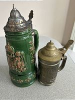 Photo showing two beer steins side by side on a table, one large green ceramic with pewter lid and one smaller metal with brass lid