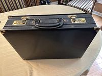 Closed black leather briefcase with gold-tone combination locks, corner protectors, and leather handle, resting on a table