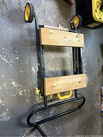 Folded view of black metal frame miter saw stand with two rectangular wooden boards and yellow wheels and handle.