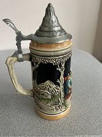 Side view of the German beer stein showing colorful raised relief of a man in traditional Bavarian attire and mountain scenery. Pewter lid with embossed design and thumb lever visible.