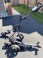One golf push cart stood upright next to a folded identical golf push cart, showing the frame, wheels, and handle.
