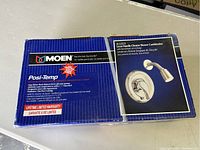 Front view of Moen Posi-Temp shower faucet box showing product branding, features including pressure balance technology, and chrome finish showerhead and lever handle.
