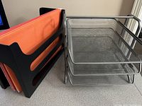 Black vertical file organizer holding orange file folders alongside silver metal mesh three-tier file tray on desk surface.