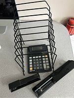 Photo showing black metal file rack, Casio calculator, black stapler, and black hole punch on a gray surface