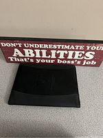 Photo of office sign with text: 'DON'T UNDERESTIMATE YOUR ABILITIES That's your boss's job.' and closed black notepad holder.