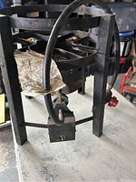 Front angle close-up photo showing metal frame, propane hose, and control valve with red knob attached to the burner base. A paper tag is also attached to the burner.