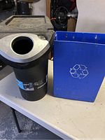 Black plastic stackable recycling bin with round opening for cans and blue metal recycling bin with white recycling symbol for paper, arranged on table.