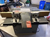 Front view of the Delta Router Shaper showing metal table surface, clamps, and central chute with warning stickers.