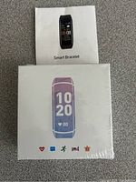Photo showing sealed white box packaging for the smart fitness bracelet with color display image, fitness and health icons, and a printed user manual placed above the box.
