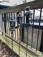 Three black metal tiki-style patio lanterns with stakes for outdoor installation shown attached to wooden railing.