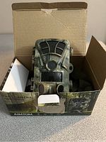 Trail camera inside its box with camouflage design and strap visible on side.