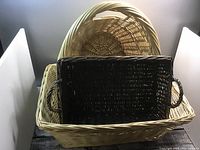 Three woven baskets stacked: small dark brown rectangular basket with handles placed inside large light beige rectangular basket, which sits on large light beige oval basket with taller sides.