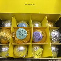 Box containing 12 assorted metallic-wrapped Laguna Moon bath bombs in gold, silver, and purple foil with scent labels.