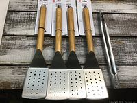 Four stainless steel spatulas with wooden handles and one pair of stainless steel tongs arranged side by side on rustic wooden surface