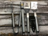 Four BBQ utensils arranged on a wooden surface: pair of stainless steel tongs, basting brush with metal handle and yellow bristles, two stainless steel slotted spatulas, and a black grill cleaning brush.