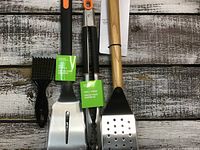 Four BBQ utensils laid out on a wooden surface; includes basting brush, spatula, tongs, and slotted spatula