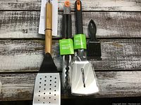 Photo of four BBQ utensils: wooden-handled slotted spatula, stainless steel tongs, stainless steel spatula, black bristle brush.