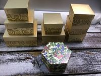 View of 10 decorative gift boxes arranged on wooden surface. Includes 8 gold-tone rectangular boxes with swirl pattern and 2 hexagonal floral print boxes.